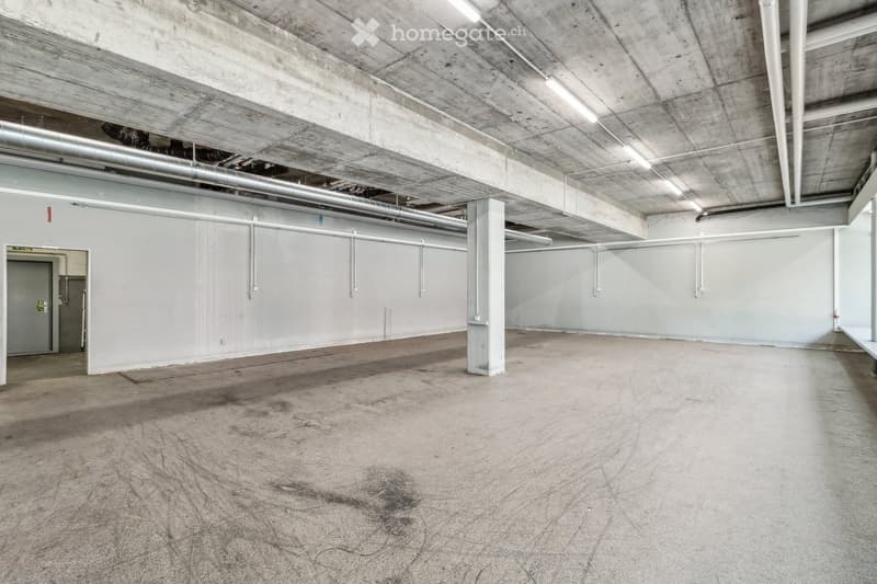 Empty warehouse with white walls, concrete floor, and visible pipes and beams on the ceiling.