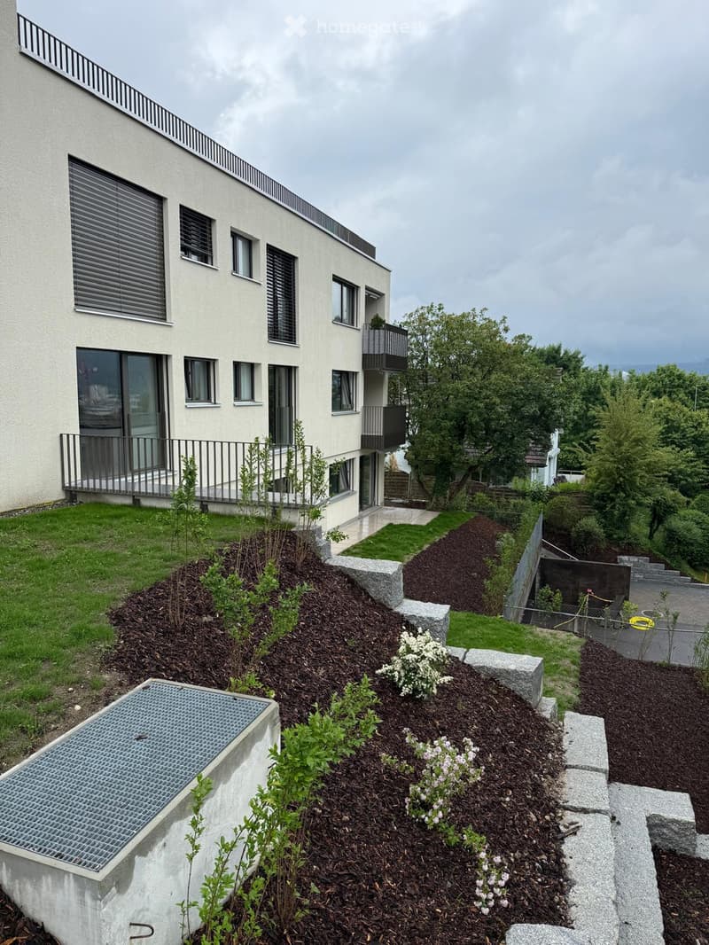 Apartment building with white facade, glass sliding doors, balconies, stairs leading to the entrance, small garden with stone steps and plants