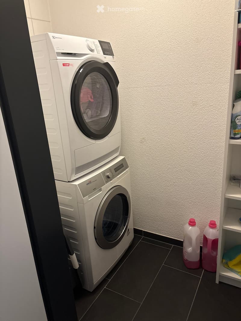 A small bathroom with a stacked washing machine and dishwasher, a white shelf with cleaning supplies, and dark tiled flooring.