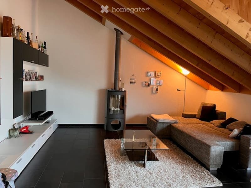 Modern living room with exposed wooden beams, a grey sectional sofa, a glass coffee table, a fireplace, and built-in shelving with decorative items.