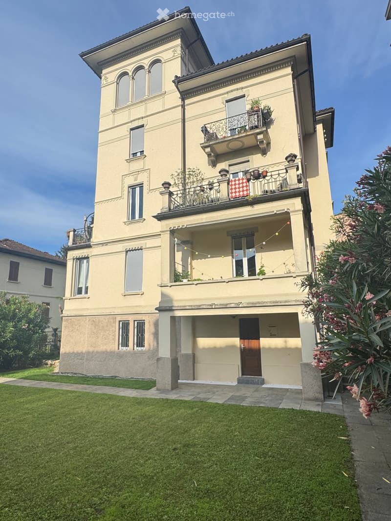 A three-story house with beige walls, multiple windows, balconies, and a well-maintained lawn in front.