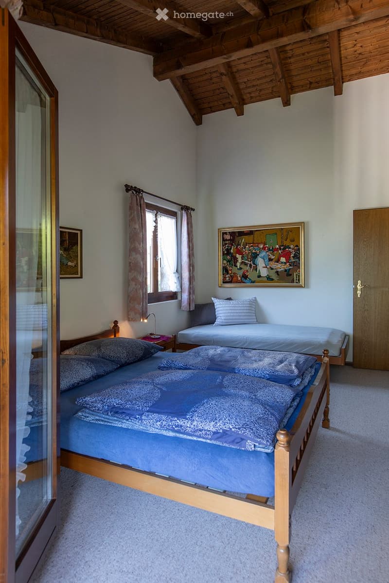 Bedroom with wooden bed, blue bedding, bedside table with lamp, small daybed, framed painting, curtains, side mirror, carpet