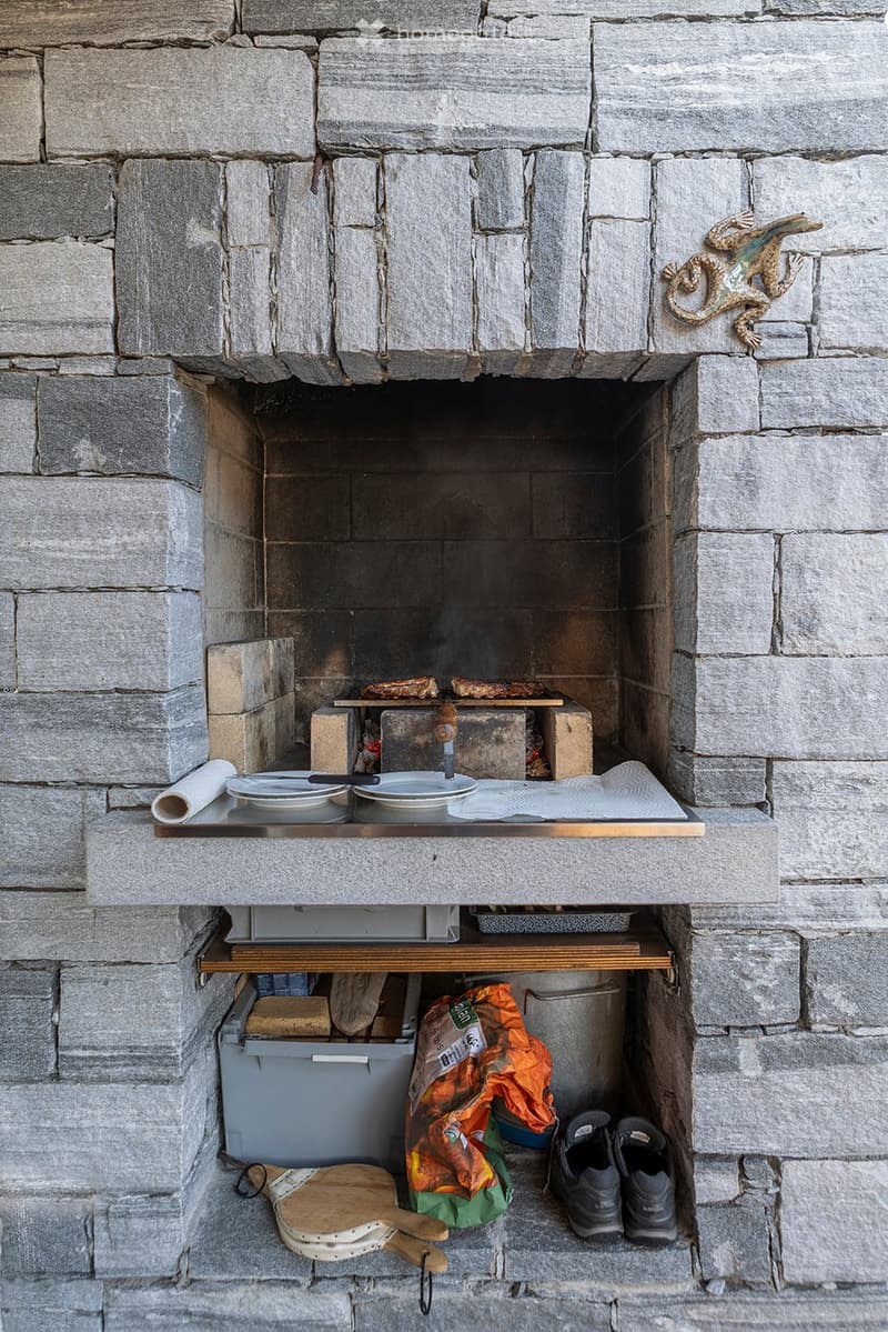 A stone fireplace with an open flame, surrounded by a hearth, with plates, a roll of paper, a basket, wooden paddles, and a bag