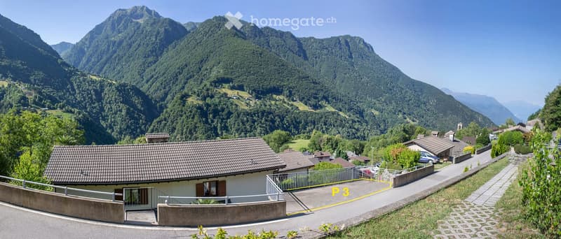 House with brown roof, surrounded by mountains, paved driveway, garden, and parking spot.