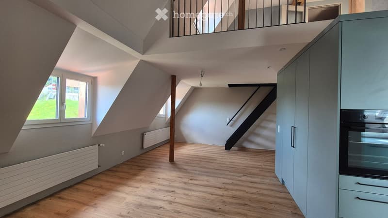 Bright, open attic space with wooden flooring, white walls, large windows, and built-in cabinets. Stairs lead to an upper level with a balcony.