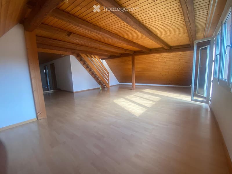 Empty room with wooden floor and exposed wooden beams on the ceiling. There is a staircase leading to another level and large windows letting in natural light.