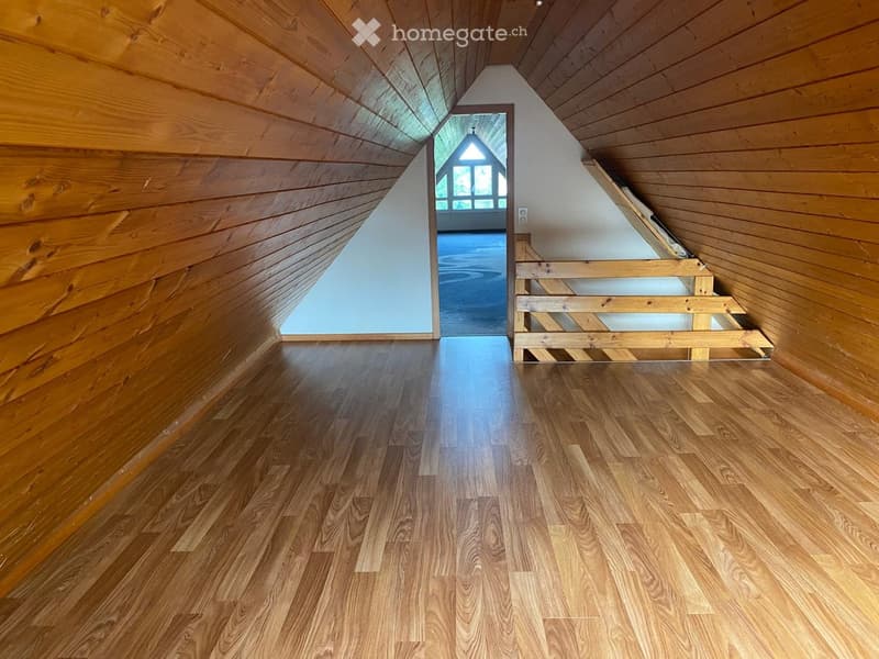 An empty attic space with wooden walls and floors, featuring a staircase leading to another room with large windows.