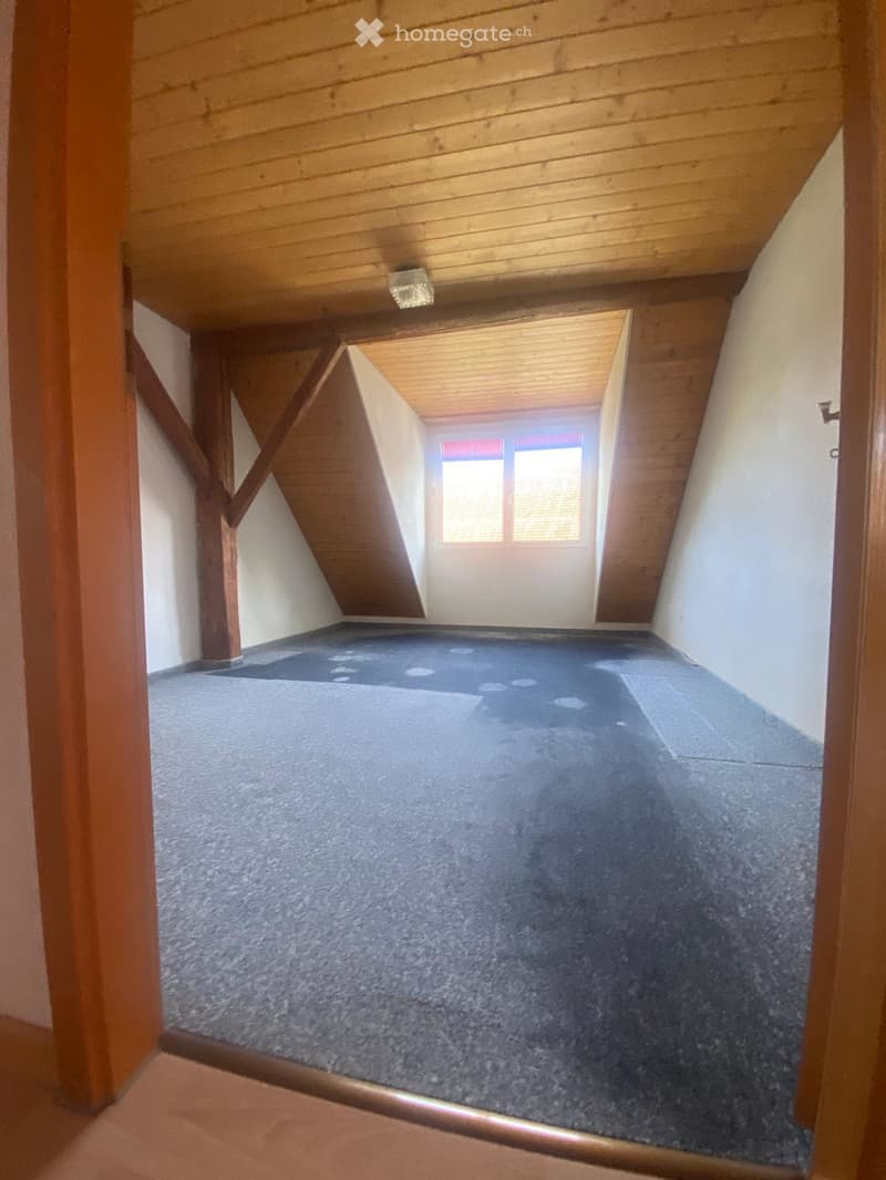 Empty attic space with wooden beams, carpeted floor, and a small window letting in natural light.