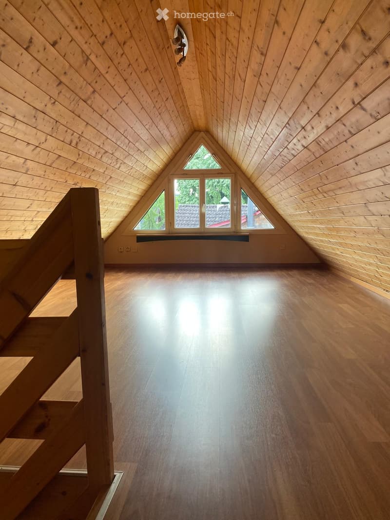 Bright, empty attic space with wooden walls, ceiling, and floor, featuring large triangular windows and a staircase leading up.