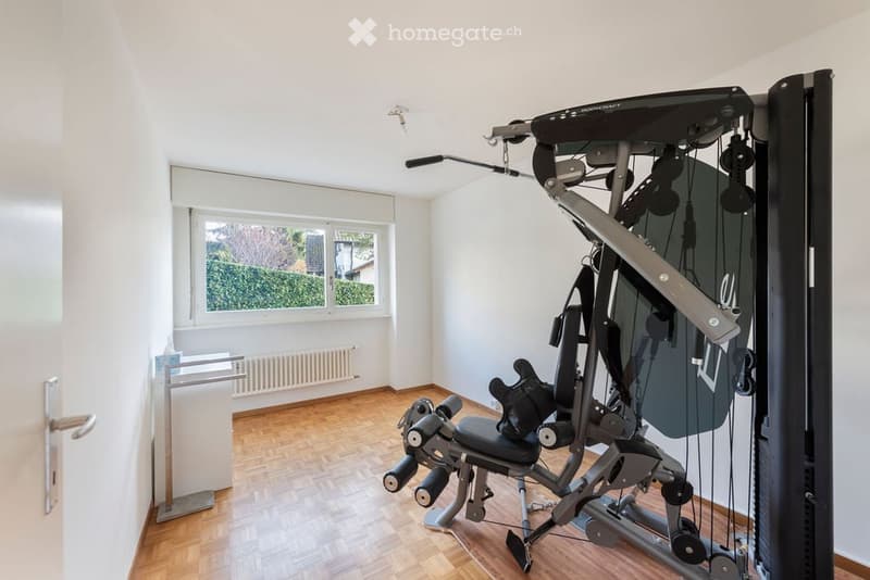 Furnished Room. A well-lit room with wooden flooring and white walls, featuring a multi-functional exercise machine, a door, a radiator, and a large window with a view of greenery.