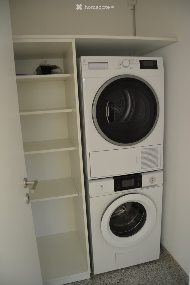 A small laundry area with a white washing machine and dryer stacked vertically, next to an empty white cabinet with multiple shelves.