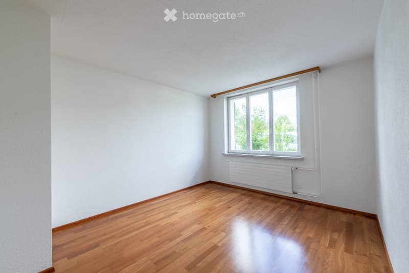 Empty room with wooden flooring, white walls, and a single window with white frame and radiator below it.