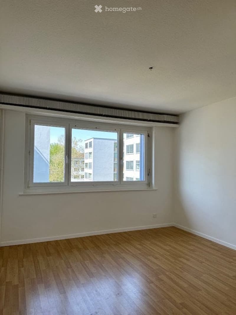 Empty room with wooden flooring, white walls, and a large window offering a view of neighboring buildings.