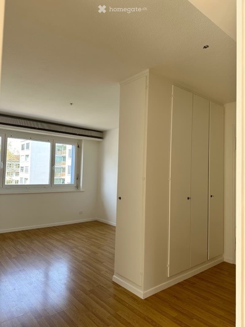 Empty room with wooden flooring, white walls, and a large built-in wardrobe. A window offers a view of the outside buildings.