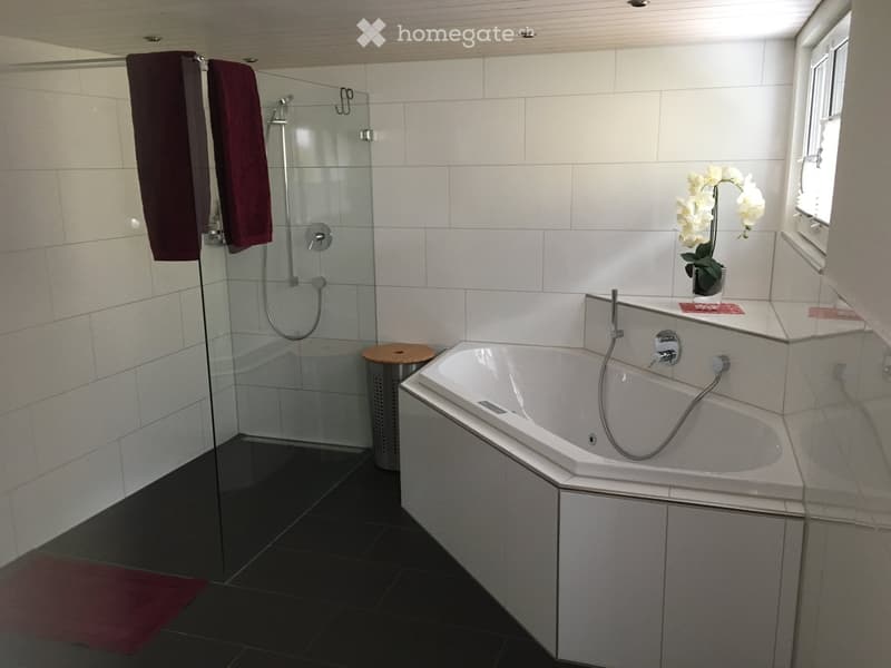 Modern bathroom with a white bathtub, glass-enclosed shower, dark tiled floor, and white tiled walls. Includes a towel rack with maroon towels, a small flower vase on the windowsill, and a trash can.