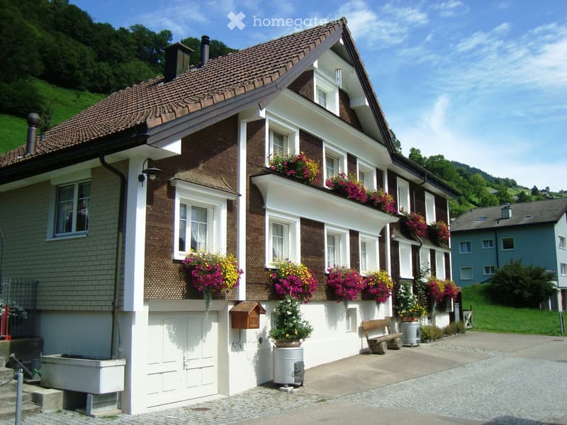 Two-story house with brown tiled roof, white trim, multiple windows adorned with colorful flower boxes, and a small garden area with a bench and potted plants.