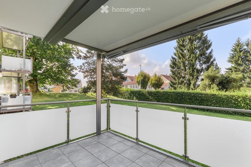 Modern balcony with glass balustrade, tiled floor, and a view of a green garden and neighboring houses.