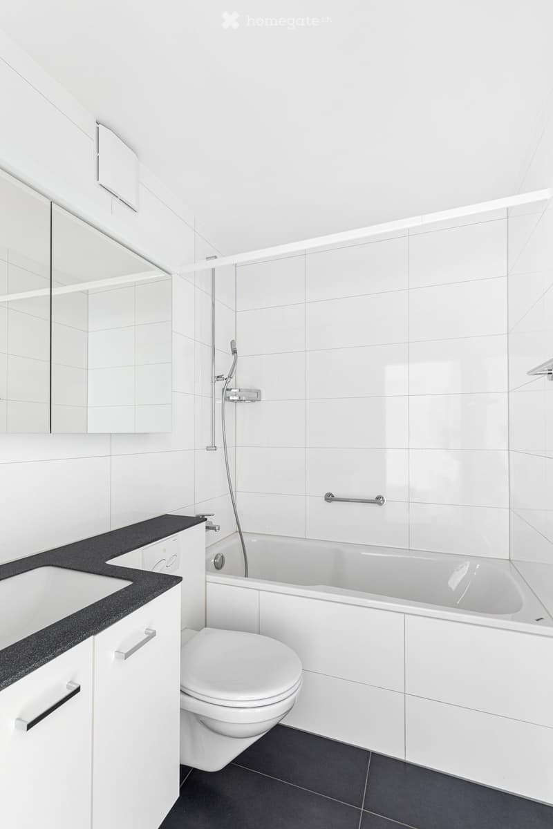 Modern bathroom with white tiles, a bathtub, toilet, sink, and mirrored cabinet.