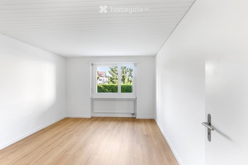 Empty room with white walls, wooden flooring, and a window with a view of greenery and a house outside.