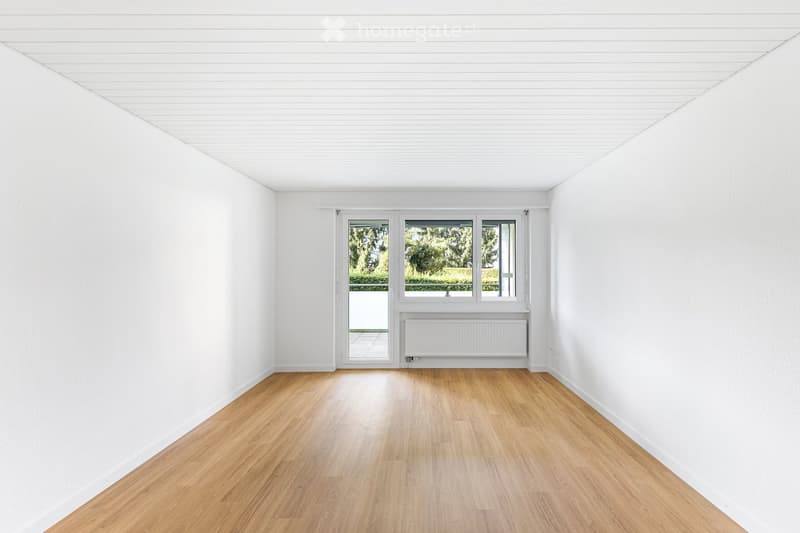 Empty room with white walls, wooden floor, large window, and radiator. No furniture or equipment present.