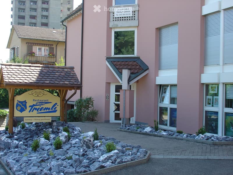 2 story pink building with white window frames, gravel entrance with sign, building in back with balconies and red flowers