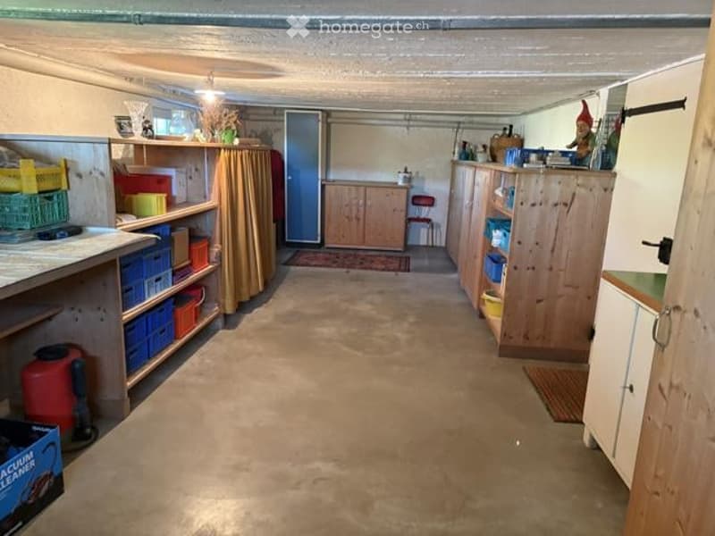 The image shows a well-organized basement or utility room with wooden shelves, cabinets, and a small table. There are various storage items, including boxes, containers, and a chair. The space has a concrete floor and is well-lit with natural light coming