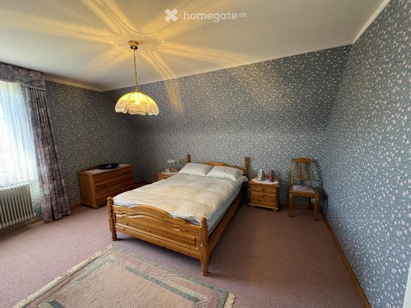 A cozy bedroom with patterned wallpaper, a wooden bed with white bedding, a nightstand, a wooden dresser, a chair, and a hanging lamp.