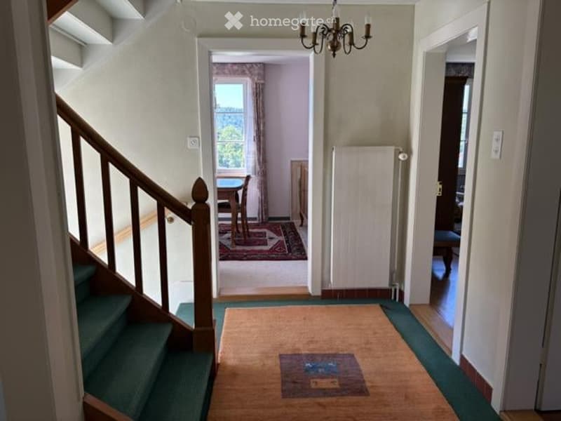 Hallway with wooden flooring, green carpeted stairs, chandelier, and visible rooms including a dining area with a table and chairs.