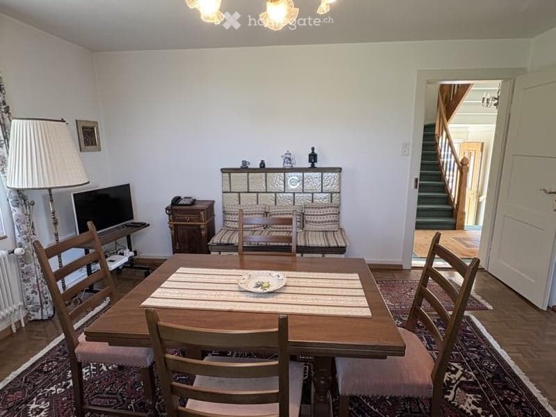 A cozy dining and living area with a wooden dining table, chairs, a sofa, a television, and a floor lamp. The room features a patterned rug and a staircase leading to another level.