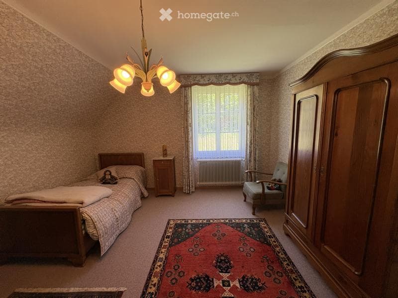 Cozy bedroom with a wooden bed, nightstand, armchair, large wardrobe, patterned area rug, and a chandelier with multiple light bulbs.