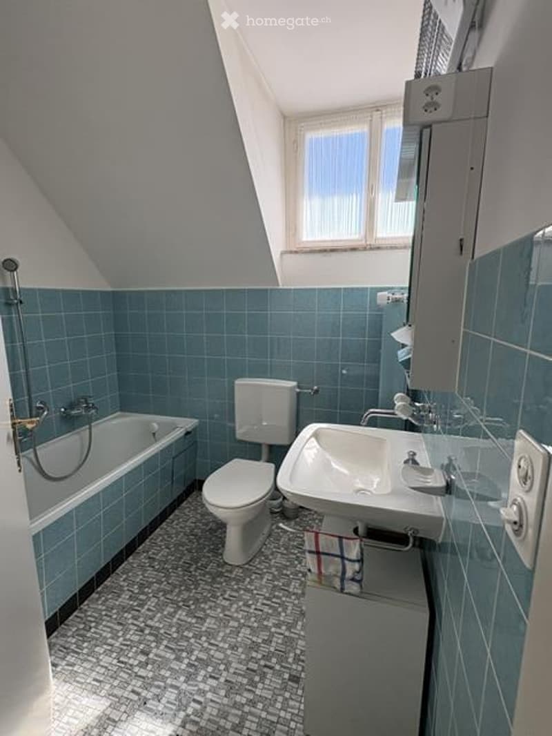 A small, cozy bathroom with blue tiled walls and a patterned mosaic floor. Features include a bathtub, toilet, sink, and a window letting in natural light.