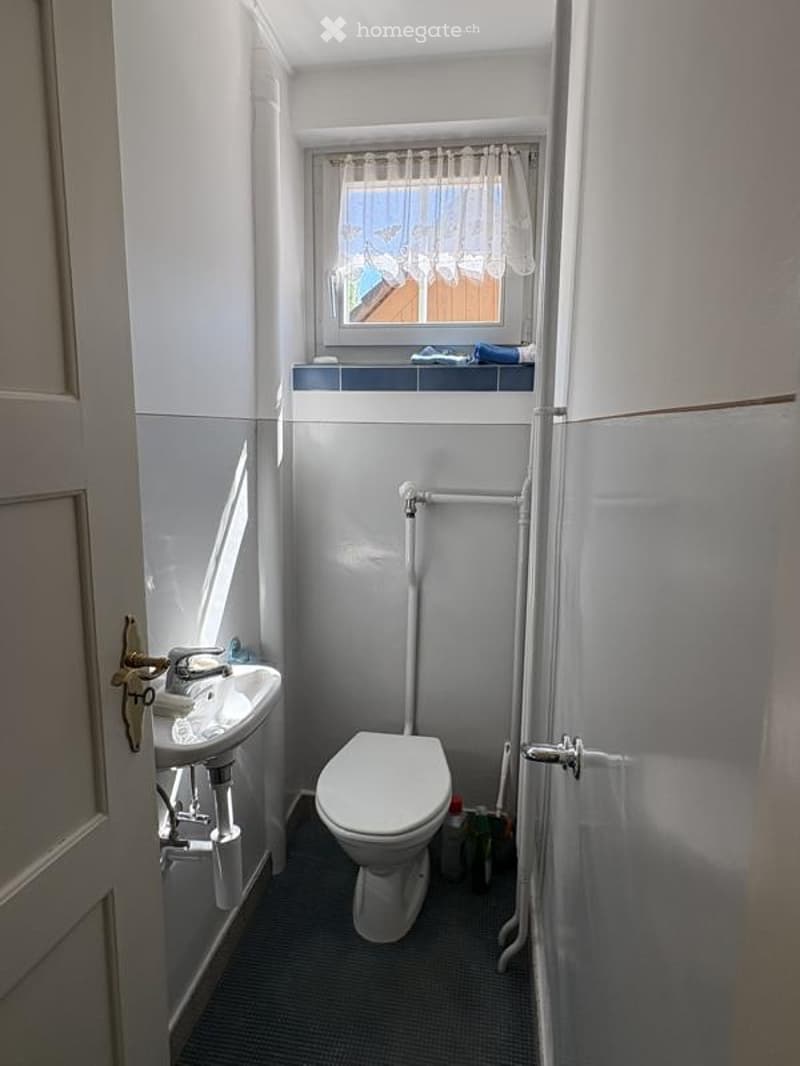 Small bathroom with a toilet, sink, and window with a curtain. The walls are white, and the floor is dark.