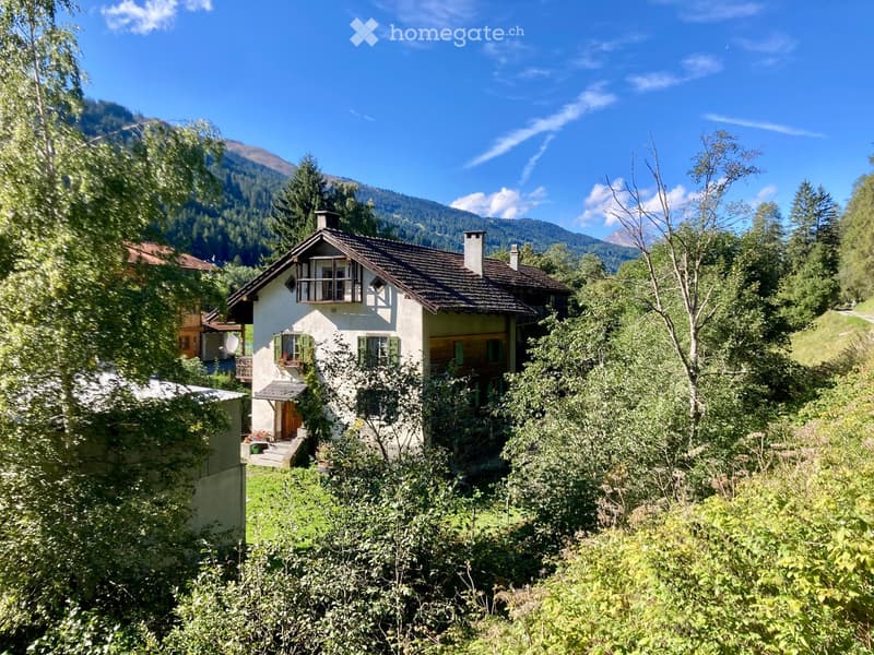 A two-story house with a tiled roof, white exterior, green shutters, and a small balcony overlooking a lush, green landscape with mountains in the background.