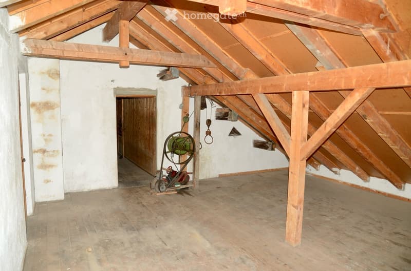Empty attic space with wooden beams, exposed wooden floor, and a small doorway leading to another room. There is a small mechanical device with a wheel and a pulley system in the center.