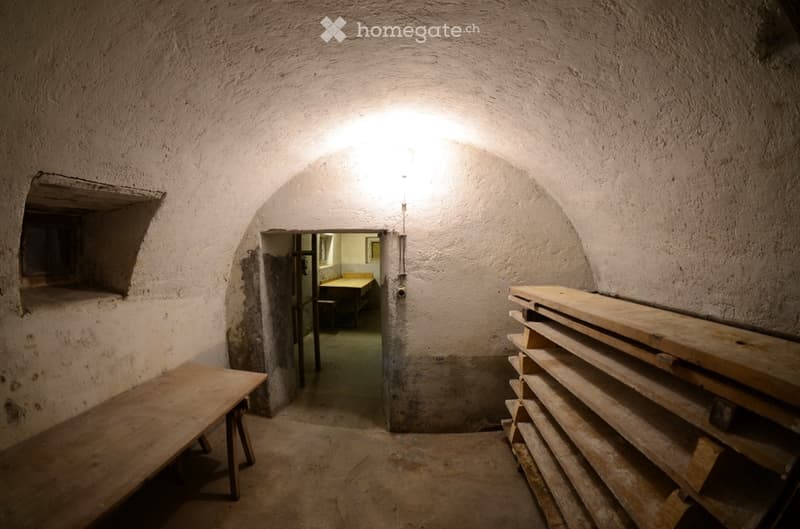A narrow, arched stone corridor with rough, textured walls and a single wooden bench on the left side. A doorway leads to another room with a table and chairs visible in the background.