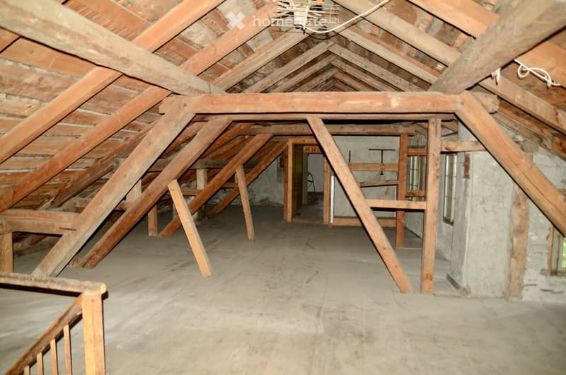 Empty attic space with exposed wooden beams and concrete floor, featuring a doorway leading to another area.