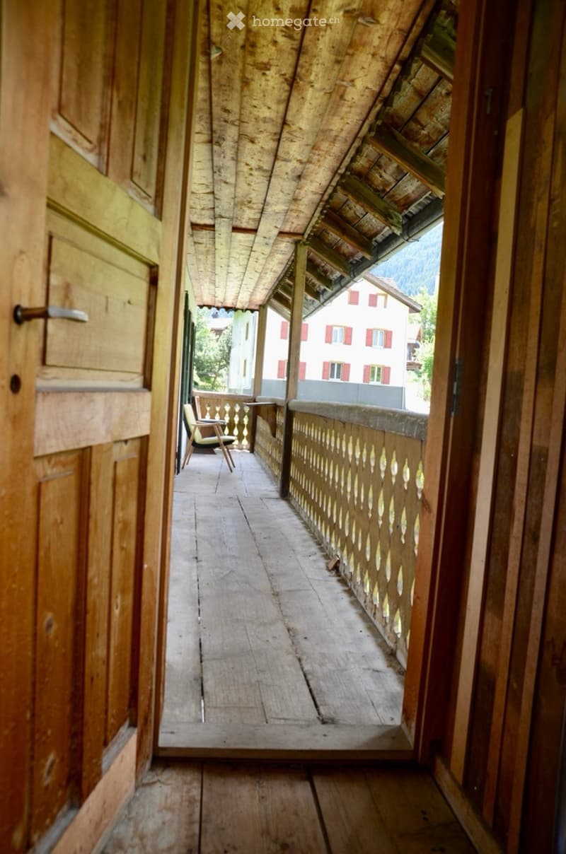 Wooden balcony with traditional wooden railing, wooden floor, and a view of a house with red shutters in the background.