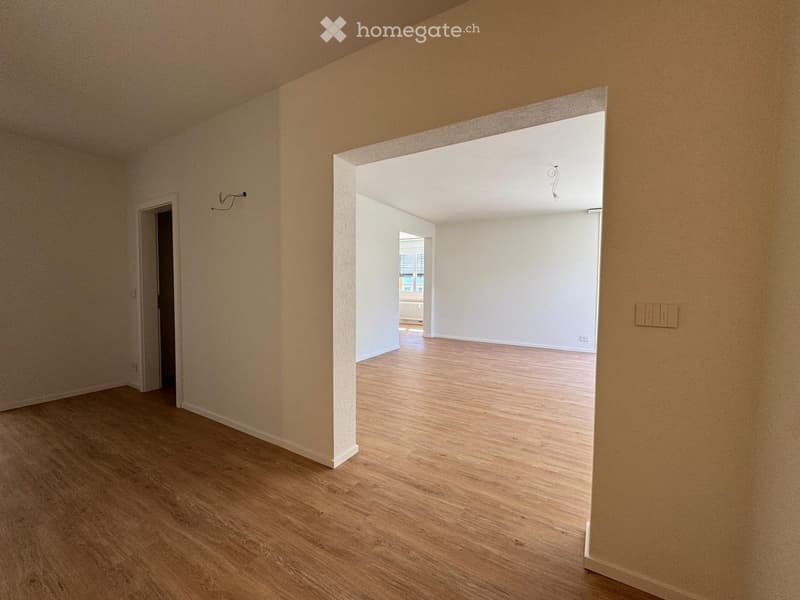 Empty room with light wooden flooring and beige walls, featuring an open doorway leading to another room with white walls and a window.