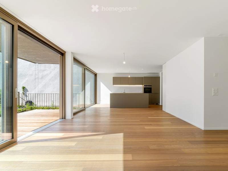 Open-plan space with wooden flooring, large glass sliding doors leading to a balcony, modern kitchen area with island and cabinets.