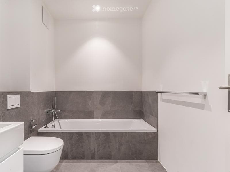 Modern bathroom with a white bathtub, toilet, and sink, featuring grey tiled walls and a sleek chrome towel rack.