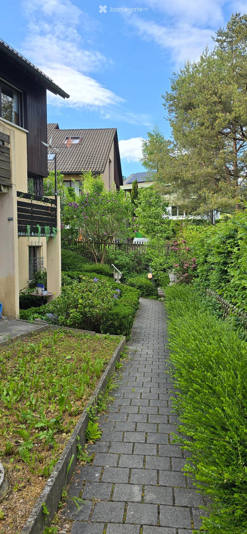 A narrow, paved pathway lined with lush greenery and shrubs leads to a small garden area with a table and chairs, surrounded by residential buildings.