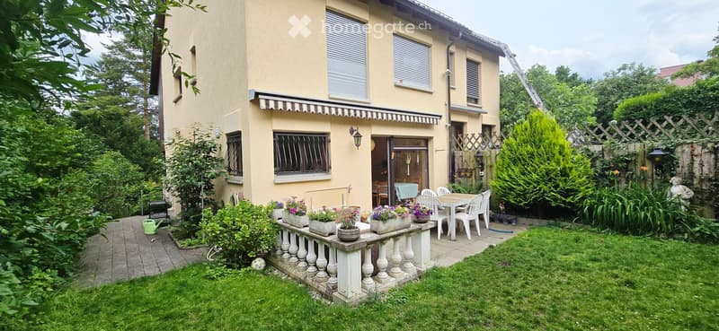 A well-maintained garden with a raised patio area, white balustrade, outdoor furniture, potted plants, and a small lawn.
