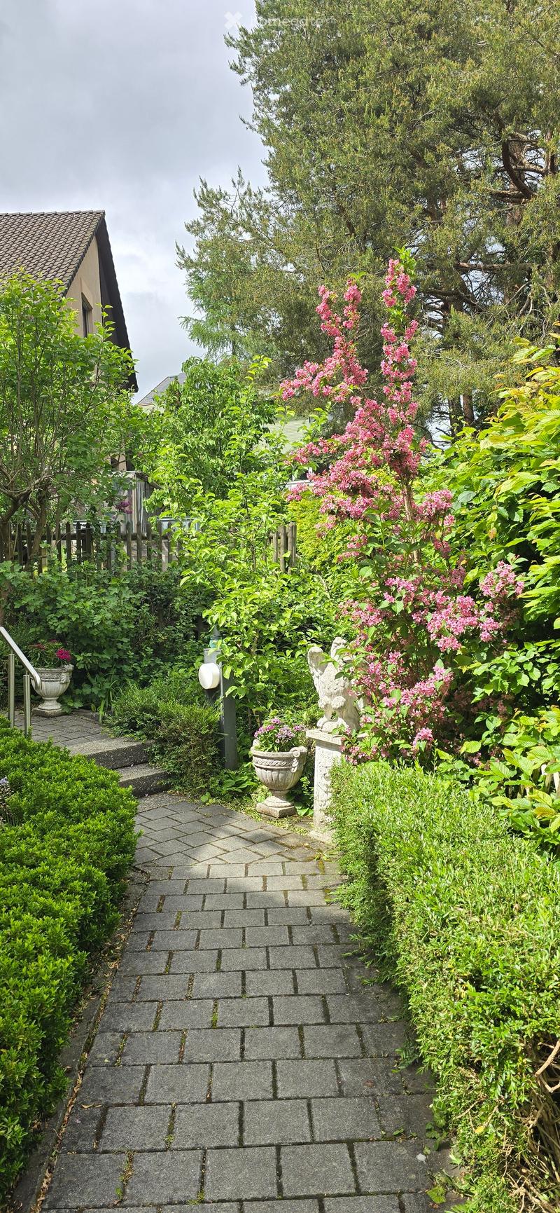 A well-maintained garden with a brick pathway, lush greenery, flowering shrubs, and decorative statues.