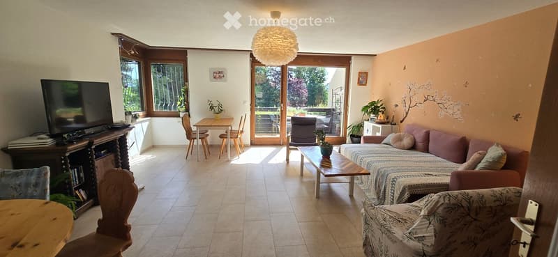 Bright living room with tiled floor, large windows, and a sliding glass door leading to a balcony. Features a patterned sofa, coffee table, dining table with chairs, and a television on a wooden stand.