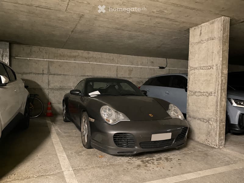 Underground parking area with several cars, including a black Porsche, a silver car, and a bicycle, with concrete pillars and a concrete ceiling.