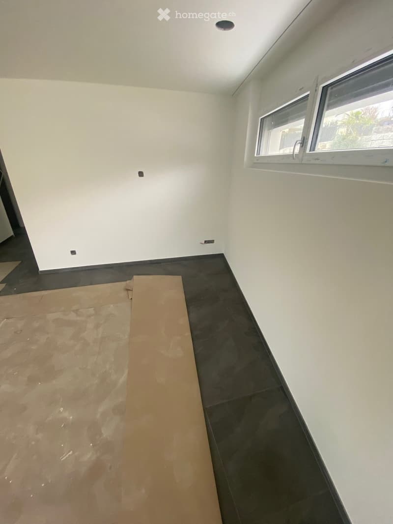 Empty room with white walls, dark tiled floor, and a single window with white frame. A piece of cardboard is placed on the floor.