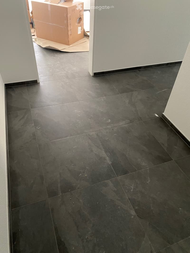 Empty room with dark grey tiled flooring and light grey walls. A cardboard box is visible in an adjacent room.