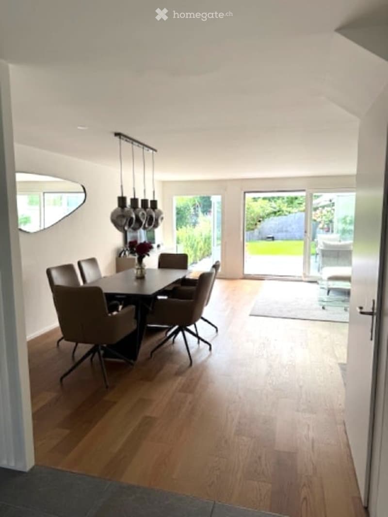 Bright dining room with wooden flooring, a black dining table surrounded by six chairs, pendant lights above, and a large mirror on the wall. A sliding glass door leads to a garden view.