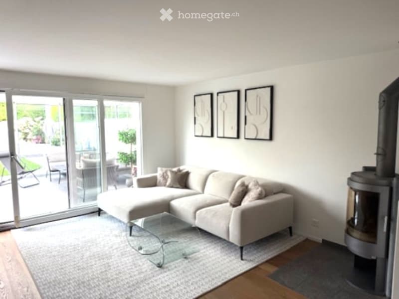 Modern living room with large windows, a sectional sofa, a glass coffee table, and a fireplace. The room features a neutral color palette with light-colored furniture and artwork on the walls.