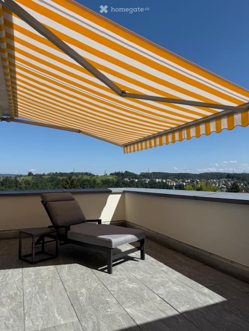 Balcony with awning, lounge chair, table, tiled floor, distant view of trees and houses